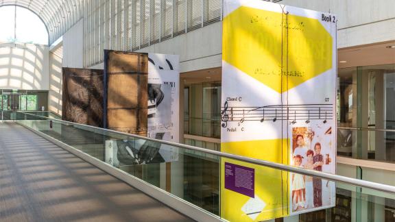 Three colorful banners hang beside a catwalk in a light-filled galleria.