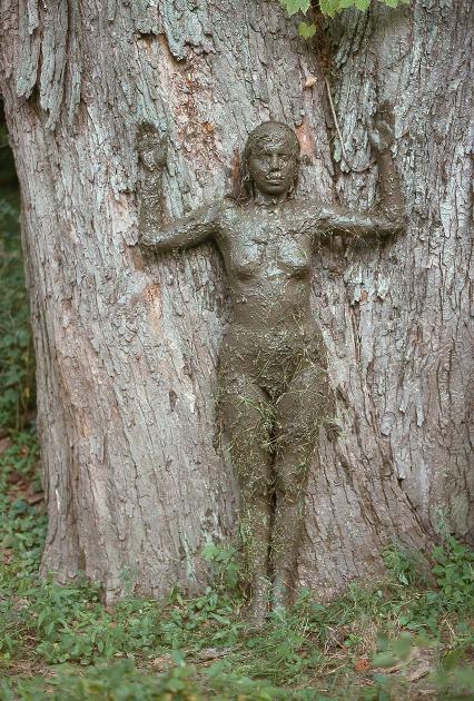 A woman covered in mud stands with her arms up and back against a tree.