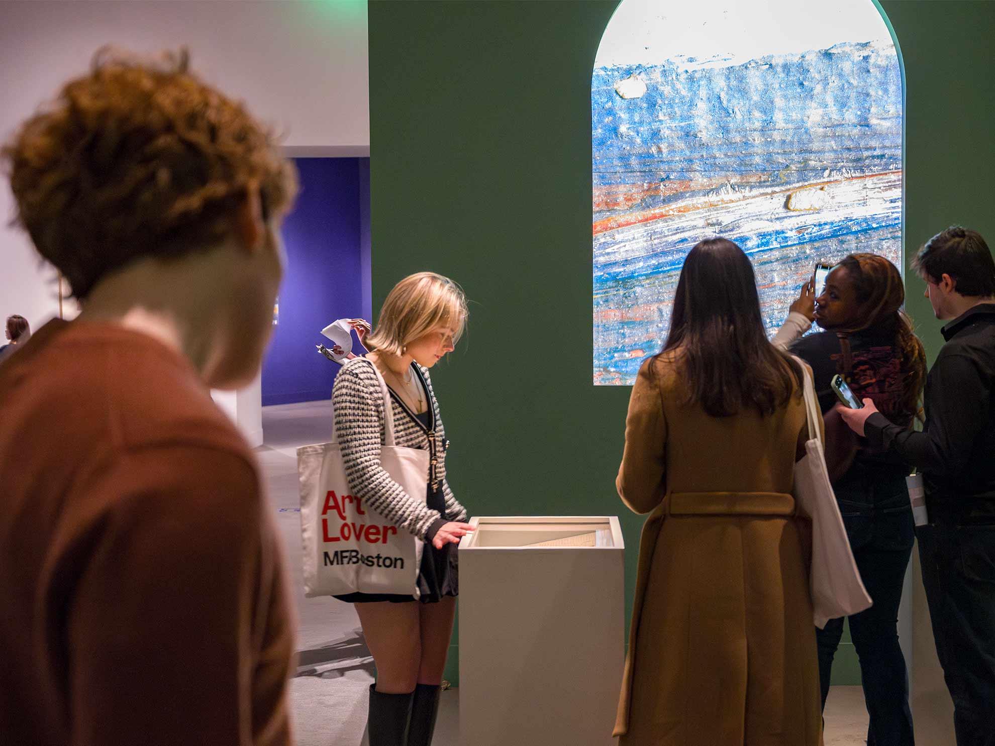 Visitor with Art Lover tote bag looking down into display case with artwork embedded within; three visitors to the right taking photos of digital projection of strokes of color