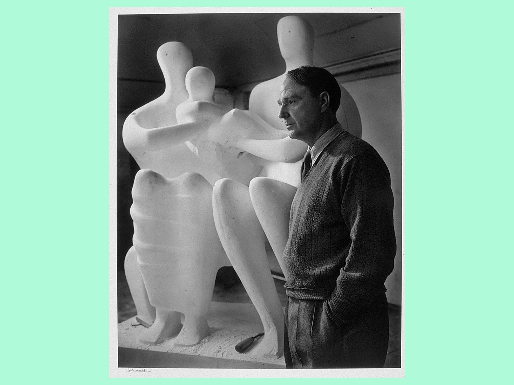 Black-and-white photograph of Henry Moore wearing a sweater) standing in front of one of his sculptures in the background