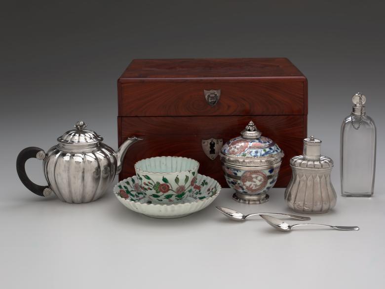 A photo of a tea service consisting of a teapot; tea canister; sugar bowl and cover; tea bowl and saucer; scent flask; two teaspoons; fitted wooden box with silver hardware