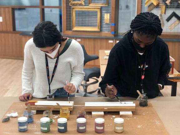 Two teens standing at work table and applying paint to pieces of a frame using paintbrushes