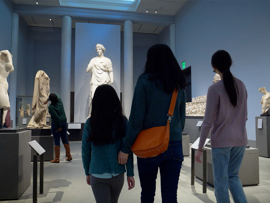 Parent and two children walking through Gods and Goddesses gallery featuring many ancient Greek sculptures on display atop pedestals