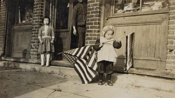 Black and white photograph of little girl holding two flags, another young girl and an adult male behind her
