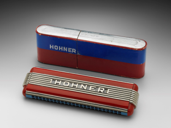 A gray and red harmonica sits in front of a blue, red, and gray case, both with the word "Hohner" on them, against a gray backdrop.